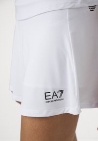 Bela atletska kratak krilo iz gladkega materiala, z vezanim črnim logotipom "EA7 Emporio Armani" na robu. Kratek, modernen kroj.