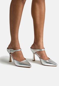 Mule argentate, metalliche, con tacco alto, punta affusolata, cinturino alla caviglia con hardware decorativo e una texture liscia e sinuosa.