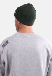 Un sweat gris avec un petit patch logo noir et or sur l'épaule gauche, associé à un bonnet tricoté vert foncé. Tissu texturé visible.