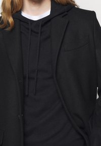 Schwarzer Blazer aus Wollmischung mit Reverskragen über einem schwarzen Kapuzenpullover mit Kordeln, was einen Layering-Look schafft.