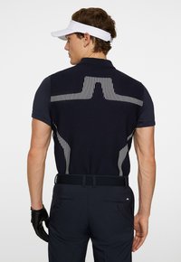 Marinblå polo med randig textur på baksidan, i kombination med mörk marinblå byxor. Utrustad med en vit skärm och svarta handskar.