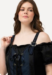 Donna con pelle chiara e capelli castano ondulati, che indossa un top nero senza spalle e un corsetto blu scuro con fibbie decorative e nastri.