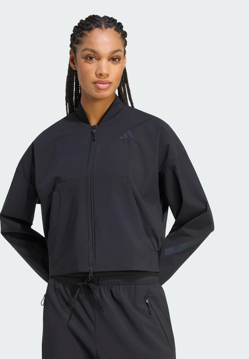 Femme aux cheveux tressés portant une veste de sport noire zippée et un pantalon à cordon assorti sur un fond uni.