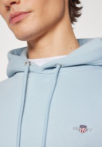 Light blue hoodie featuring a drawstring hood, silver hardware, and an embroidered logo on the chest. Soft texture and casual design.