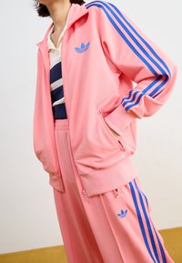 Rosa träningsoverall med blå tre-stripes-accenter. Jackan har dragkedja och fickor, kompletterad med matchande byxor och logotypdetalj.