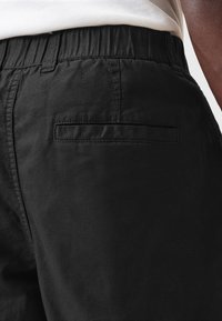 Svarta shorts med elastisk midja, som har en platt bakficka och en slät, texturerad tyg. Närbild av midjeområdet.