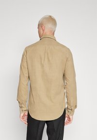 Chemise à manches longues de couleur beige clair, en tissu texturé avec un ourlet courbé et des poignets à boutons, vue de dos.