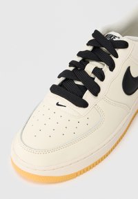 Sneaker de cor creme com atacadores pretos, apresentando um cabedal de couro liso, bico perfurado e uma sola de borracha texturizada em cor de borracha.