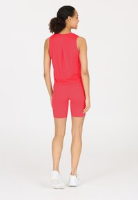 Rode sportieve tanktop met een plooi aan de achterkant, gecombineerd met bijpassende rode shorts. Model draagt witte sneakers. Glad, rekbaar materiaal.