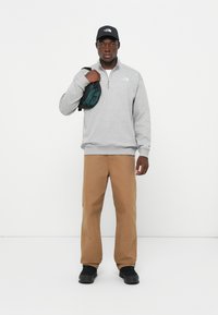 Pullover grigio in felpa con zip, pantaloni beige, cappellino nero e una piccola borsa verde e nera. Le scarpe nere completano l'outfit.