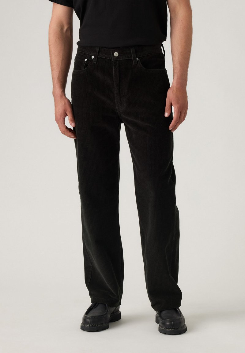 Man in zwarte corduroy broek, zwarte schoenen en een zwart shirt, staande tegen een effen lichte achtergrond.