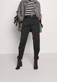 Pantalones cargo negros con bolsillos laterales, combinados con un abrigo gris de pata de gallo con detalles de flecos y una camiseta de rayas en blanco y negro.