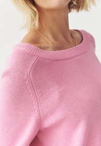 Růžový pletený svetr s širokým výstřihem, ribbed lemy a jemným texturovaným stehem podél švů. Měkký, lehký materiál.