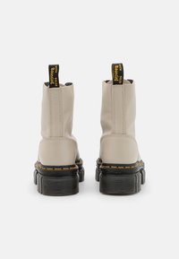 Botas de tobillo beige con un acabado brillante, que cuentan con suelas de goma negra, costuras amarillas y una lengüeta trasera con texto. Diseño de suela texturizada.