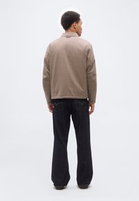 Άνδρας που στέκεται με την πλάτη γυρισμένη, φορώντας μπεζ σακάκι και σκούρα μπλε τζιν παντελόνι με φαρδιά μπατζάκια, μπροστά σε μονόχρωμο φωτεινό φόντο.