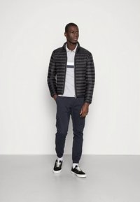 Veste matelassée noire avec piqûres horizontales, polo gris avec une bande blanche et bleue, pantalon cargo bleu marine et baskets noires avec semelles blanches.