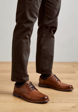 Brown leather shoes with black laces, round toes, and textured sides. Positioned on light-colored wooden flooring with dark trousers.