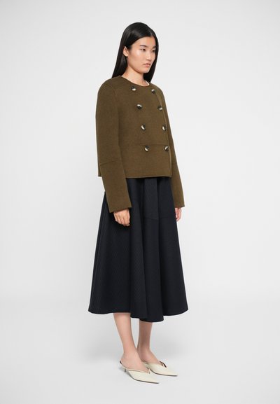 Oliivroheline kahekordne jakk suurte mustade nuppudega, kombineeritud voolava musta seelikuga, millel on vertikaalsed triibud ja kreemjad libistatavad kingad.