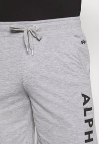 Pantalon de survêtement gris avec une taille à cordon de serrage, des poches latérales et un grand logo noir "ALPHA" imprimé verticalement sur la jambe gauche. Texture douce.
