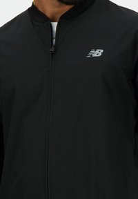 Zwarte ritssluiting jacket met een ribbelkraag, een gladde structuur en een zilveren New Balance-logo op de linkerborst.