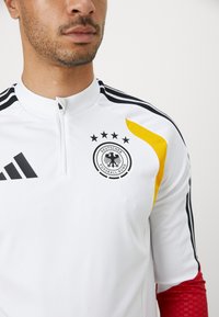 Λευκή αθλητική φανέλα με ψηλό γιακά, μαύρες ρίγες, κίτρινες λεπτομέρειες και το λογότυπο της Γερμανικής Ποδοσφαιρικής Ομοσπονδίας με πέντε αστέρια.