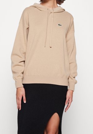 Beżowa bluza z kapturem i sznurkami oraz małym zielonym logo krokodyla, noszona z czarną prążkowaną spódnicą z przednim rozcięciem.