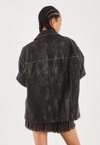 Personne avec des cheveux tressés portant une veste en cuir noir et marron surdimensionnée et une jupe plissée sombre, de dos contre un fond blanc.
