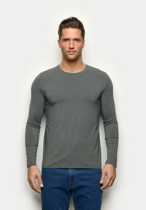 Mann mit kurzen dunklen Haaren, der ein eng anliegendes langärmliges graues Hemd und blaue Jeans trägt, vor einem schlichten hellen Hintergrund stehend.