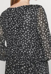 Kvinna i en svart klänning med genomskinliga långa ärmar, dekorerad med små vita blommönster, visad bakifrån från midjan och uppåt.