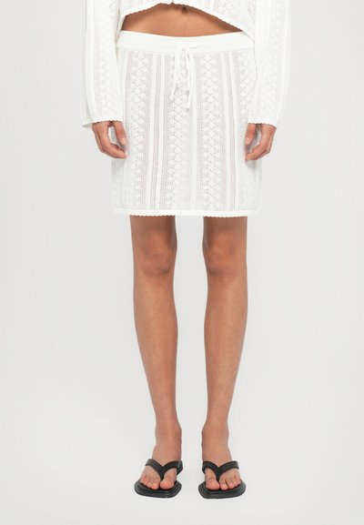 Jupe mini blanche tricotée avec une taille à cordon de serrage, présentant un motif texturé et un ourlet en forme de vagues. Assortie à des tongs noires.