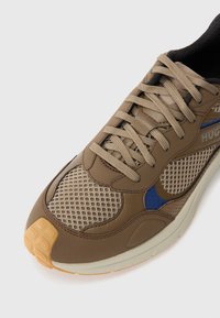 Brown athletic shoe featuring mesh panels, beige accents, and a textured rubber sole. Laces in matching color and small blue detailing visible.