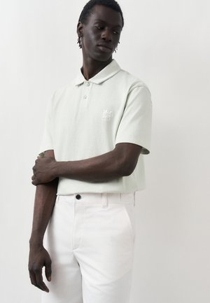 Άνδρας που φοράει ανοιχτό γκρι κοντομάνικη πόλο με λογότυπο και λευκό παντελόνι, στέκεται μπροστά σε απλό ανοιχτό φόντο με σταυρωμένα χέρια.