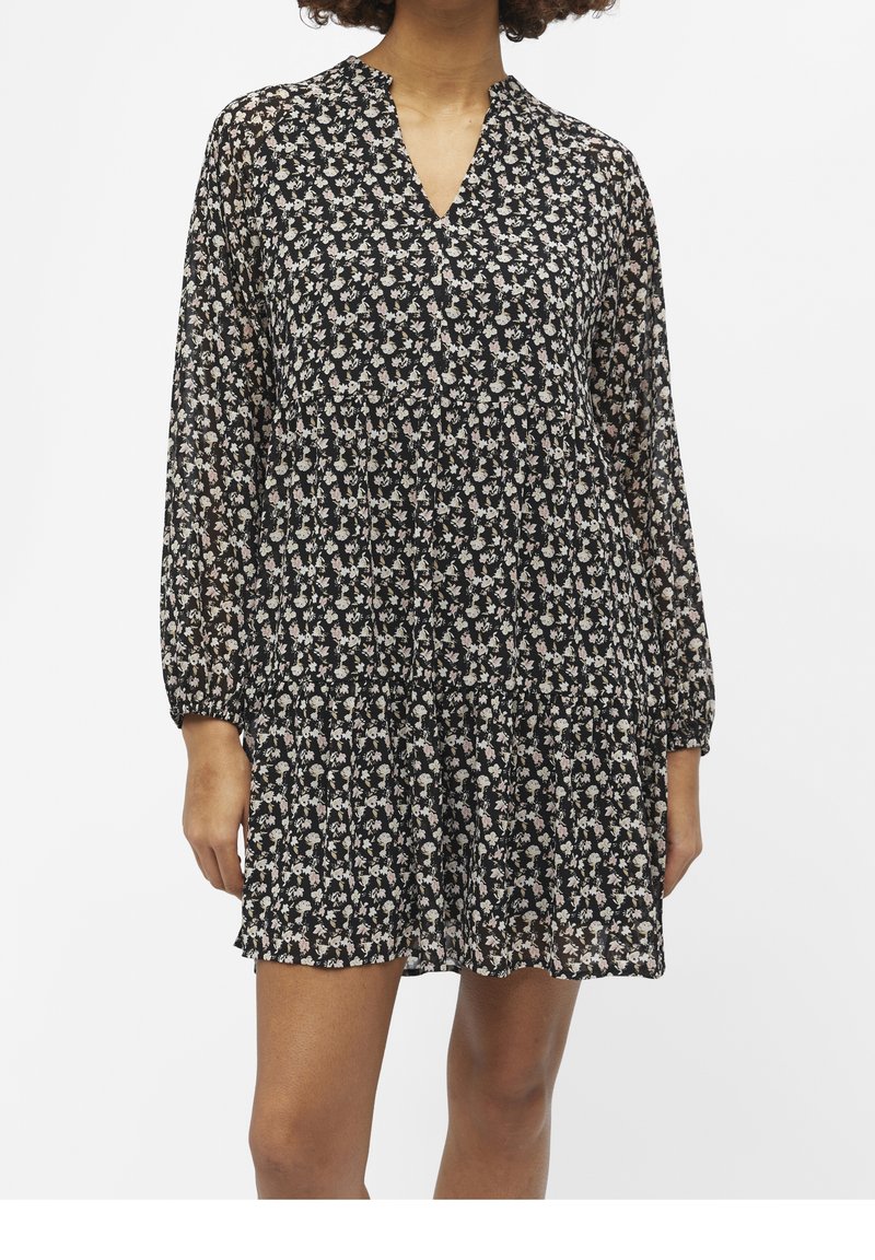 Robe florale noire avec un décolleté en V, des manches longues et transparentes, et une silhouette fluide. Présente de petits motifs de fleurs blanches et roses partout.