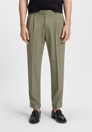 Pantaloni verde oliva con una vestibilità sartoriale, pieghe frontali e chiusura a bottoni, abbinati a mocassini neri e risvolti.