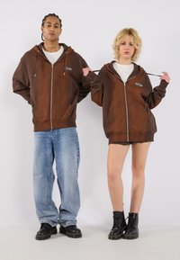 Brown zip-up hoodies with front pockets, worn over white shirts. The male model in baggy jeans; the female model wears a mini skirt and black boots.