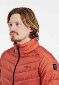 Giacca puffer rossa con colletto alto, chiusura a zip e dettagli neri. Presenta una texture trapuntata diagonale e un logo del marchio sul petto e sulla manica.