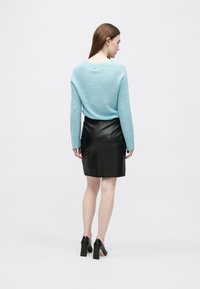 Žena s rovnými hnedými vlasmi, oblečená v svetlomodrom svetri, čiernej koženej sukni a čiernych lodičkách, otočená chrbtom na bielom pozadí.