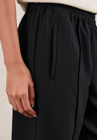 Čierne joggers nohavice vyrobené z hladkej tkaniny, s elastickým pásom a bočnou vreckom. Detailný záber ruky položené na vrecku.