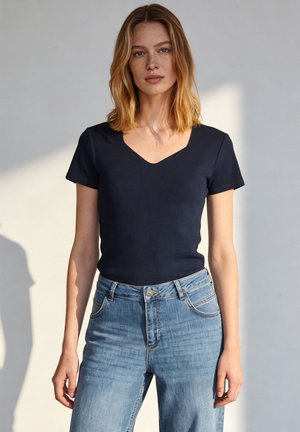 Junge Frau mit schulterlangem blondem Haar, die ein schwarzes V-Ausschnitt-Kurzarmshirt und blaue Jeans trägt, stehend vor hellem Hintergrund.