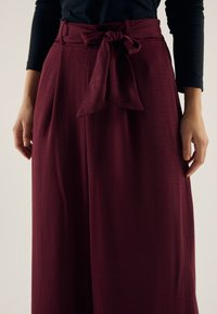 Pantalon large bordeaux en tissu texturé, avec une ceinture nouée à la taille et des plis pour plus de volume.