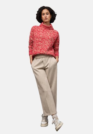 Rood en crèmekleurig geblokt coltrui, lichtbeige plooibroek en neutrale sneakers. Model staat in een ontspannen houding met de handen in de zakken.