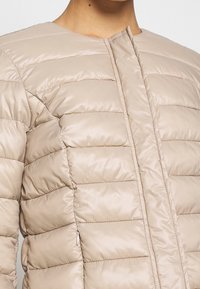 Giubbotto imbottito beige con cuciture orizzontali, colletto rotondo e apertura frontale. Il materiale sembra morbido e lucido con una superficie liscia.