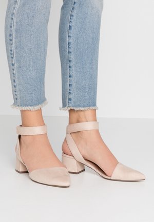 Stopy w jasnoniebieskich dżinsach z postrzępionym dołem, ubrane w beżowe zamszowe buty na niskim obcasie z zapięciem na kostkę, na gładkiej białej podłodze.