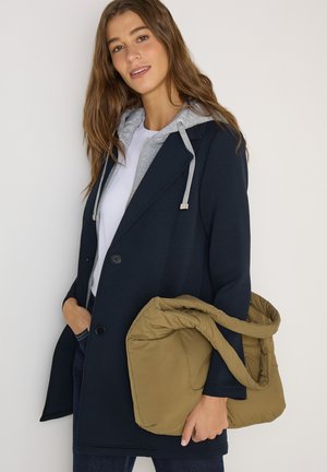 Giovane donna con indosso un cappotto blu navy e una felpa con cappuccio grigia, che tiene una grande borsa trapuntata color beige su uno sfondo bianco semplice.