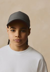 Graue Mütze mit abgerundetem Kopfteil und gebogenem Schirm, getragen über einem weißen Sweatshirt mit Rundhalsausschnitt. Der Hintergrund ist in einem neutralen Beigeton gehalten.