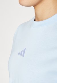 Camiseta de algodón azul claro con cuello redondo, que presenta un pequeño logo púrpura en un diseño gráfico en el pecho. Tejido texturizado.