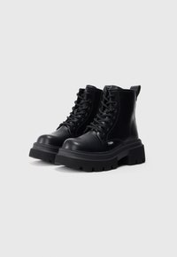Botas de cuero negro hasta el tobillo con suela de goma negra gruesa, punta redonda, diseño con cordones y una lengüeta en la parte trasera para un acceso fácil.