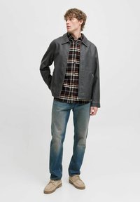 Grijze leren jas met een puntige kraag over een rood en zwart geruite shirt. Blauwe jeans en beige schoenen maken de outfit compleet. Gladde textuur, casual pasvorm.