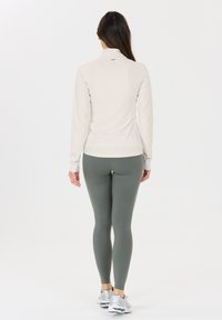 Helles graues Langarmoberteil mit hohem Kragen, kombiniert mit eng anliegenden olivgrünen Leggings, beide aus einem glatten, dehnbaren Stoff. Silberne Schuhe.