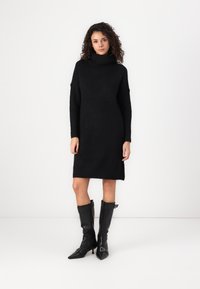 Čierne pletené šaty s vysokým zvlneným golierom, oversized rukávmi a rovnou siluetou, doplnené nadkolennými čiernymi koženými čižmami.